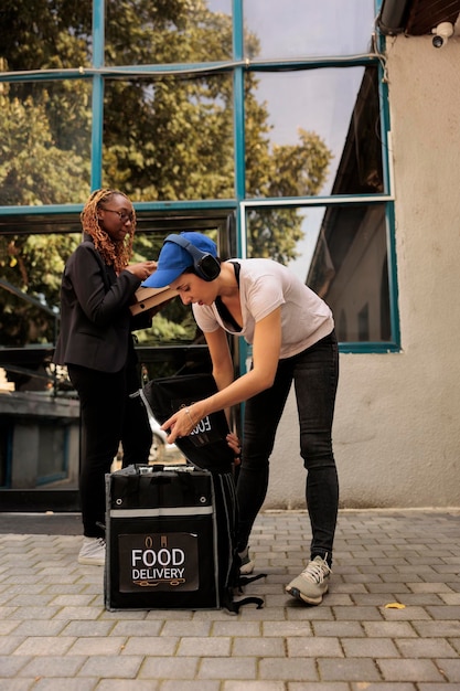 Trabajador del servicio de entrega de alimentos con auriculares sacando cajas de pizza de una bolsa térmica, el cliente recibe el pedido frente al edificio de oficinas al aire libre. Mujer entregando comida para llevar de pizzería