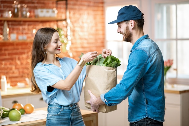 Trabajador de servicio de mensajería entregando alimentos frescos, dando bolsa de compras a una cliente mujer feliz en la cocina de casa