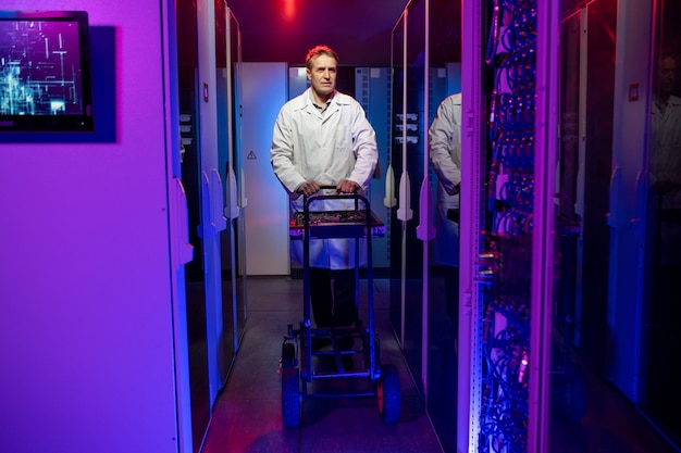 Foto trabajador de servidor maduro en bata blanca empujando el carro con herramientas mientras camina sobre el centro de datos oscuro