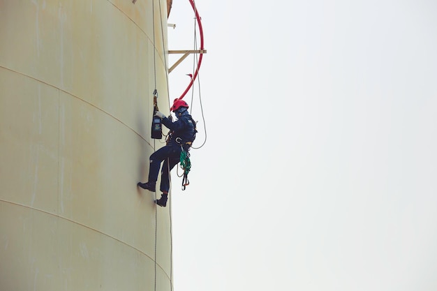 Trabajador de sexo masculino acceso industrial que trabaja en el arnés del uso del aceite del tanque de la altura, inspección del acceso de la cuerda del equipo de seguridad del casco del aceite y gas del tanque del espesor.