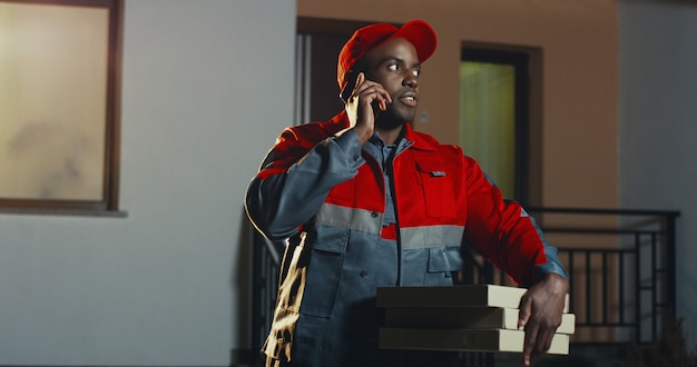 Trabajador de sexo masculino afroamericano de la entrega de pizza en el uniforme rojo y gorra de pie en la casa por la noche con cajas de pizza y hablando por el teléfono móvil. Al aire libre.