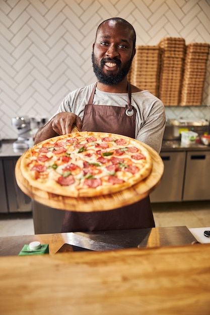 Trabajador de sexo masculino guapo en delantal mirando a la cámara y sonriendo mientras sostiene una deliciosa pizza de pepperoni