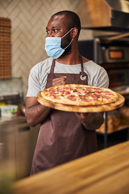 Trabajador de sexo masculino guapo en máscara médica con tablero de madera con pizza recién horneada