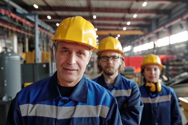 Trabajador de sexo masculino maduro de gran planta industrial o fábrica o capataz en ropa de trabajo y casco protector de pie contra dos colegas