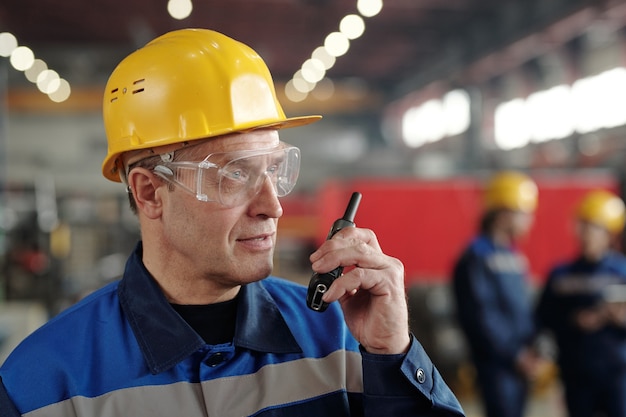 Trabajador de sexo masculino maduro de planta industrial o fábrica o capataz en ropa de trabajo y casco protector con walkie-talkie delante de la cámara