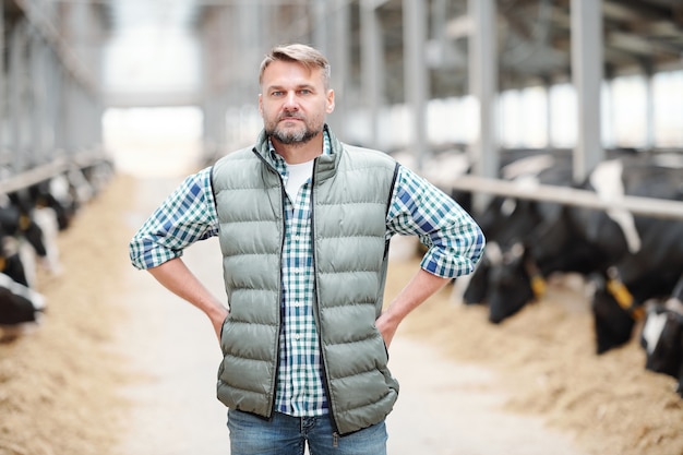 Foto trabajador de sexo masculino maduro serio o jefe de la granja lechera contemporánea de pie delante de la cámara en el fondo de largos establos