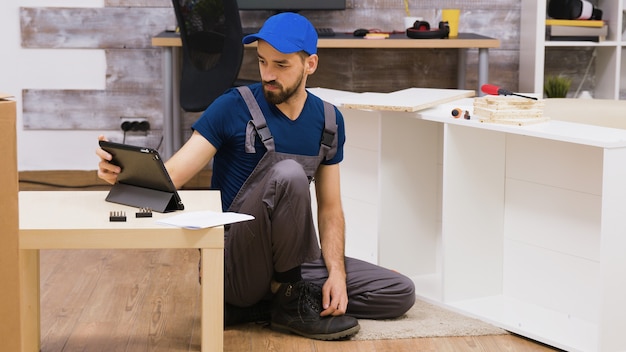 Trabajador de sexo masculino en monos con un conjunto de tapa un estante en el nuevo hogar siguiendo instrucciones de tablet PC