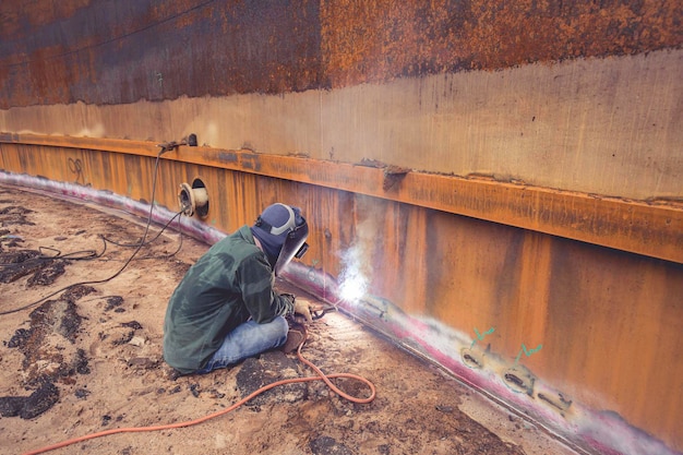 Trabajador de sexo masculino que usa ropa protectora y reparación de chispas de soldadura tanque de construcción industrial bobina aceite pesado dentro de espacios confinados