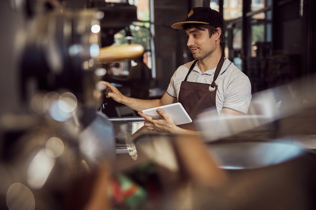 Trabajador de sexo masculino sonriente en delantal con tablet PC y equipo industrial operativo para tostar granos de café