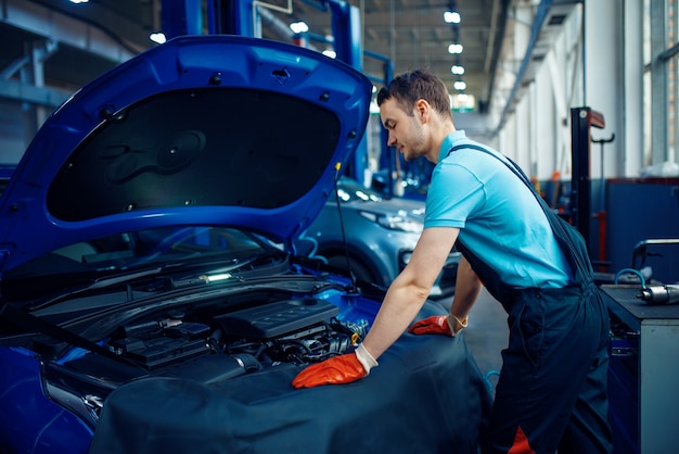 Trabajador de sexo masculino sonriente en uniforme comprueba el motor del vehículo, la estación de servicio del coche. Comprobación e inspección de automóviles, diagnóstico y reparación profesionales