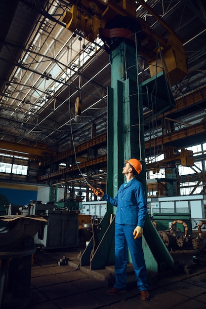 Trabajador de sexo masculino en uniforme y casco opera una grúa en la fábrica. Producción industrial, ingeniería metalúrgica, fabricación de máquinas eléctricas.