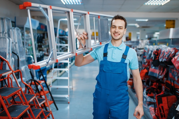 Trabajador de sexo masculino en uniforme sostiene nuevas escaleras de aluminio en almacén de herramientas. Departamento con escaleras, elección de equipo en ferretería, supermercado de instrumentos