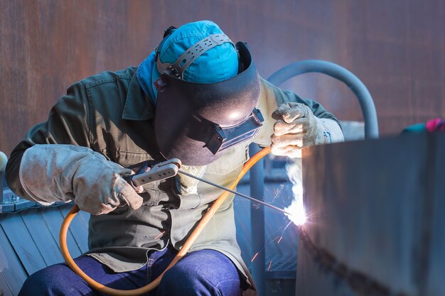 Trabajador de sexo masculino vistiendo ropa protectora y placa de reparación de soldadura de aceite de tanque de construcción industrial dentro de espacios confinados.