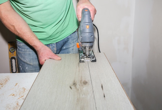 El trabajador sostiene en las manos una sierra de calar eléctrica y corta el suelo laminado en el apartamento. Mantenimiento, reparación, trabajos de renovación con sierra de calar. Tablones de parquet de madera en el interior.