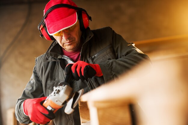 Trabajador de tela senior moderno en un uniforme profesional y protección trabajando con una amoladora eléctrica en una paleta de madera.