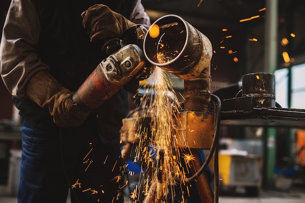 Trabajador de tela en tubo de metal de corte uniforme protector sobre la mesa de trabajo con una amoladora eléctrica en el taller industrial.