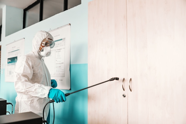 Foto trabajador en uniforme blanco estéril, con guantes de goma y máscara en la celebración de rociador con desinfectante y armario esterilizador en el aula.