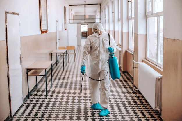Foto trabajador en uniforme blanco estéril, con máscara y gafas sosteniendo el rociador con desinfectante y rociando alrededor del pasillo en la escuela. prevención de la propagación del virus corona.