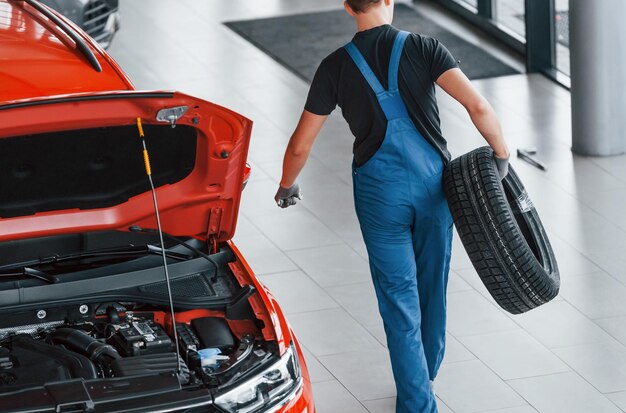 El trabajador con uniforme negro y azul sostiene la rueda del automóvil y camina con un automóvil casi rojo