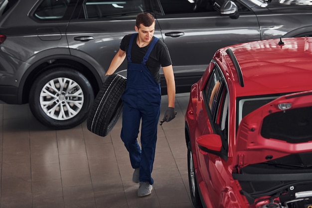Trabajador en uniforme negro y azul sostiene la rueda del coche y camina con cerca de automóvil rojo