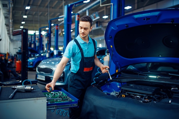 Trabajador en uniforme toma la llave de la caja de herramientas, estación de servicio de automóviles. Comprobación e inspección de automóviles, diagnóstico y reparación profesionales