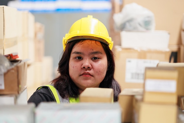 Trabajadora de almacén asiática obesa con chaleco de seguridad y casco duro en producto industrial de fábrica de almacén en cajas de cartón en estantes Mujer de talla grande inspeccionando productos mientras trabaja