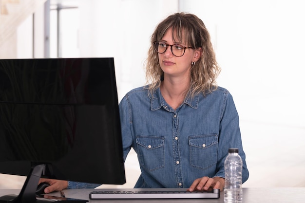 Trabajadora con camisa de jeans casual y anteojos en la oficina usando computadora en un fondo iluminado.