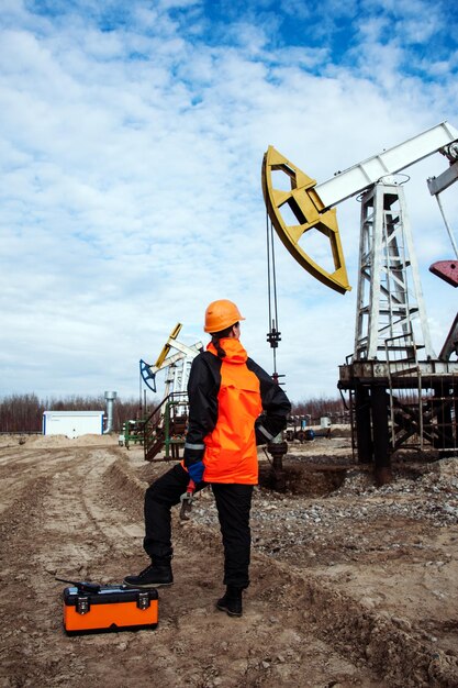 Trabajadora en el campo petrolero, con llaves en las manos, casco naranja y ropa de trabajo. Fondo de sitio industrial.