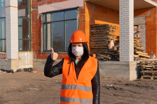 Trabajadora de la construcción en overol y máscara médica mostrando los pulgares hacia arriba en el fondo de la casa en construcción Mujer joven en casco y chaleco naranja mostrando gesto de aprobación