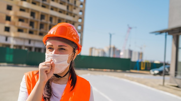 Trabajadora de la construcción con overoles y máscara médica tosiendo en el puño en el fondo de la casa en construcción Concepto de amenaza de infección epidémica por coronavirus