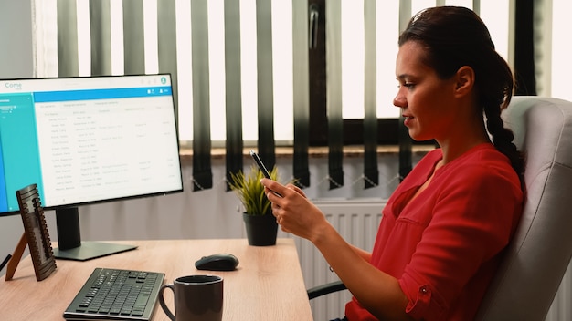 Trabajadora escribiendo en el teléfono móvil frente a la computadora en la sala de la oficina. Emprendedor hispano sentado en el lugar de trabajo de la empresa corporativa, tomando un descanso escribiendo en el teléfono inteligente frente a la computadora leyendo noticias