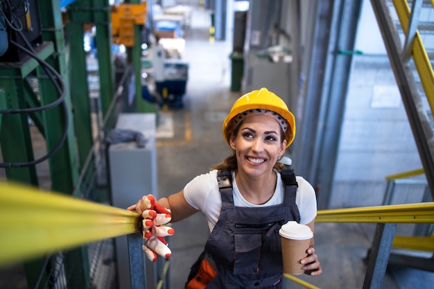 Trabajadora hermosa en uniforme y casco sosteniendo una taza de café y subiendo las escaleras de la fábrica.