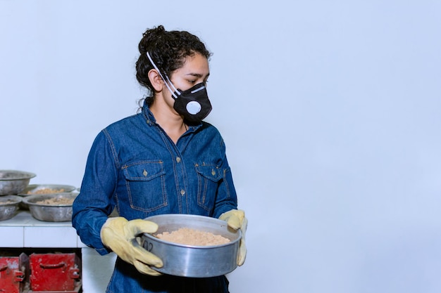 Trabajadora industrial con máscara protectora y guantes de cuero industriales sosteniendo un recipiente con arena en un laboratorio de pruebas geológicas