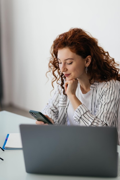 Trabajadora joven sentada en el escritorio y usando el teléfono celular