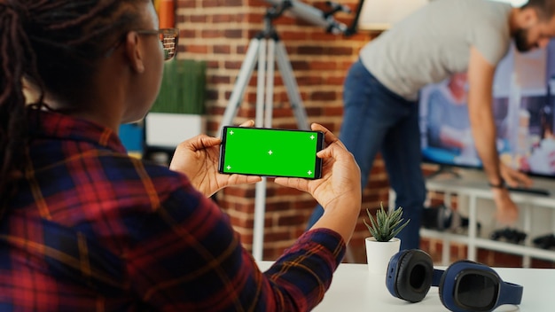 Trabajadora mirando la pantalla verde horizontal en el teléfono inteligente, usando el espacio de copia en blanco de la maqueta con la clave de croma en la pantalla. Sosteniendo el teléfono con una plantilla de espacio de copia aislada en el apartamento.