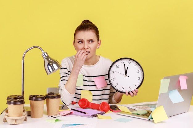 Foto trabajadora mordiéndose las uñas sosteniendo un gran reloj de pared sentado en el lugar de trabajo todo cubierto con notas adhesivas