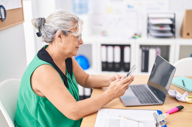 Trabajadora de negocios de cabello gris de mediana edad que usa una computadora portátil y un teléfono inteligente en la oficina