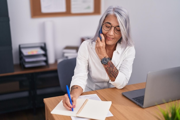 Trabajadora de negocios de mediana edad de cabello gris hablando en un teléfono inteligente escribiendo en un cuaderno en la oficina