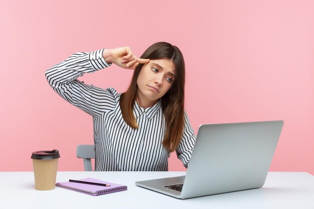 Trabajadora de oficina deprimida e infeliz con camisa a rayas sentada en el lugar de trabajo mostrando un gesto estúpido para mostrar una laptop hablando en una videollamada. Disparo de estudio interior aislado sobre fondo rosa