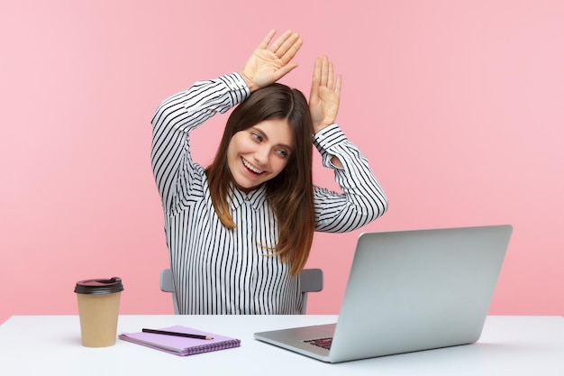 Una trabajadora de oficina juguetona que muestra orejas de conejo con las manos y sonríe mirando la pantalla de un portátil en una videollamada charlando en la cámara web y coqueteando en un estudio interior aislado en un fondo rosa