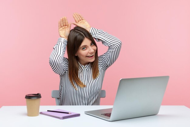 Trabajadora de oficina positiva sonriendo mostrando orejas de conejo con la mano en la cabeza descansando divirtiéndose en el lugar de trabajo en la oficina en casa sintiéndose despreocupada y feliz Foto de estudio interior aislada en fondo rosa