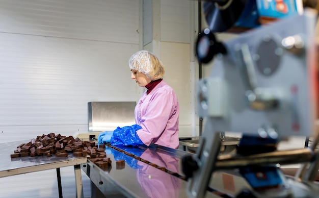 Foto trabajadora profesional en uniforme y guantes protectores clasifica dulces de chocolate en la línea de producción en la fábrica