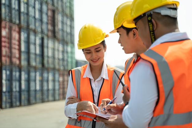 Trabajadora que trabaja con su colega, de pie con un casco amarillo para controlar la carga y verificar la calidad de los contenedores del buque de carga para importación y exportación en el astillero o puerto.