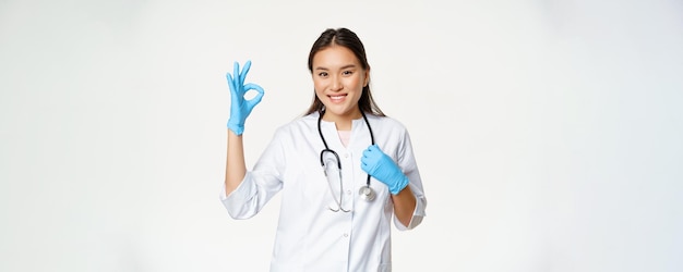 Una trabajadora sanitaria sonriente, una doctora asiática con guantes de goma y uniforme médico, muestra aprobación con un signo de fondo blanco.