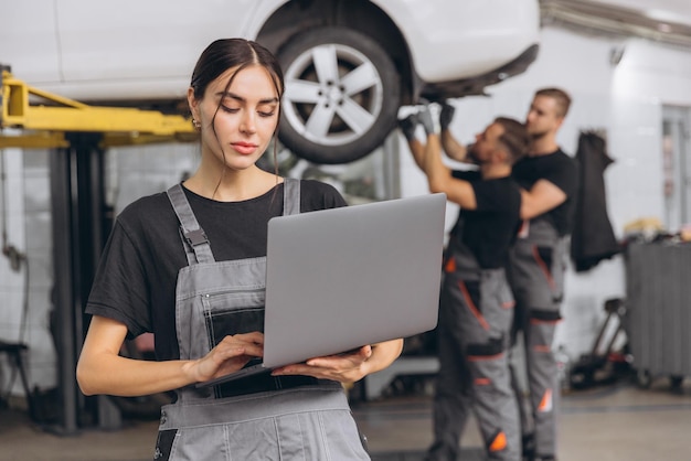 Trabajadora de servicio automovilístico hermosa mujer joven sosteniendo la computadora portátil en las manos y mirando a la cámara sonriendo mecánicos reparando automóviles levantados en el puente detrás