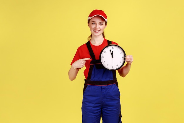 Una trabajadora sonriente y satisfecha parada y señalando un gran reloj de pared en sus manos, mirando la cámara, usando overoles y gorra roja. Disparo de estudio interior aislado sobre fondo amarillo.