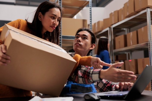 Foto trabajadores de almacenes postales que gestionan la logística de entrega en portátiles y discuten el calendario de envíos. almacén, hombre y mujer, empleados asiáticos analizando datos de distribución en la sala de almacenamiento