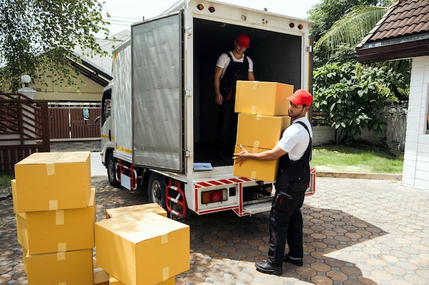 Trabajadores asiáticos y caucásicos uniformados descargando cajas de cartón del camión Repartidores descargando cajas y revisando la lista de verificación con sus compañeros de trabajo Servicio profesional de entrega y mudanza