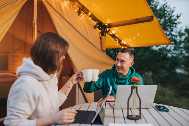 Los trabajadores autónomos de una pareja familiar feliz beben café mientras trabajan con una computadora portátil en una acogedora carpa glamping en las noches de verano Carpa de camping de lujo para vacaciones al aire libre y vacaciones Concepto de estilo de vida