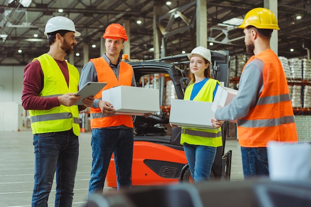 Trabajadores confiados con cascos de seguridad, ropa de trabajo, cajas en la mano, capataz hablando usando una tableta digital.